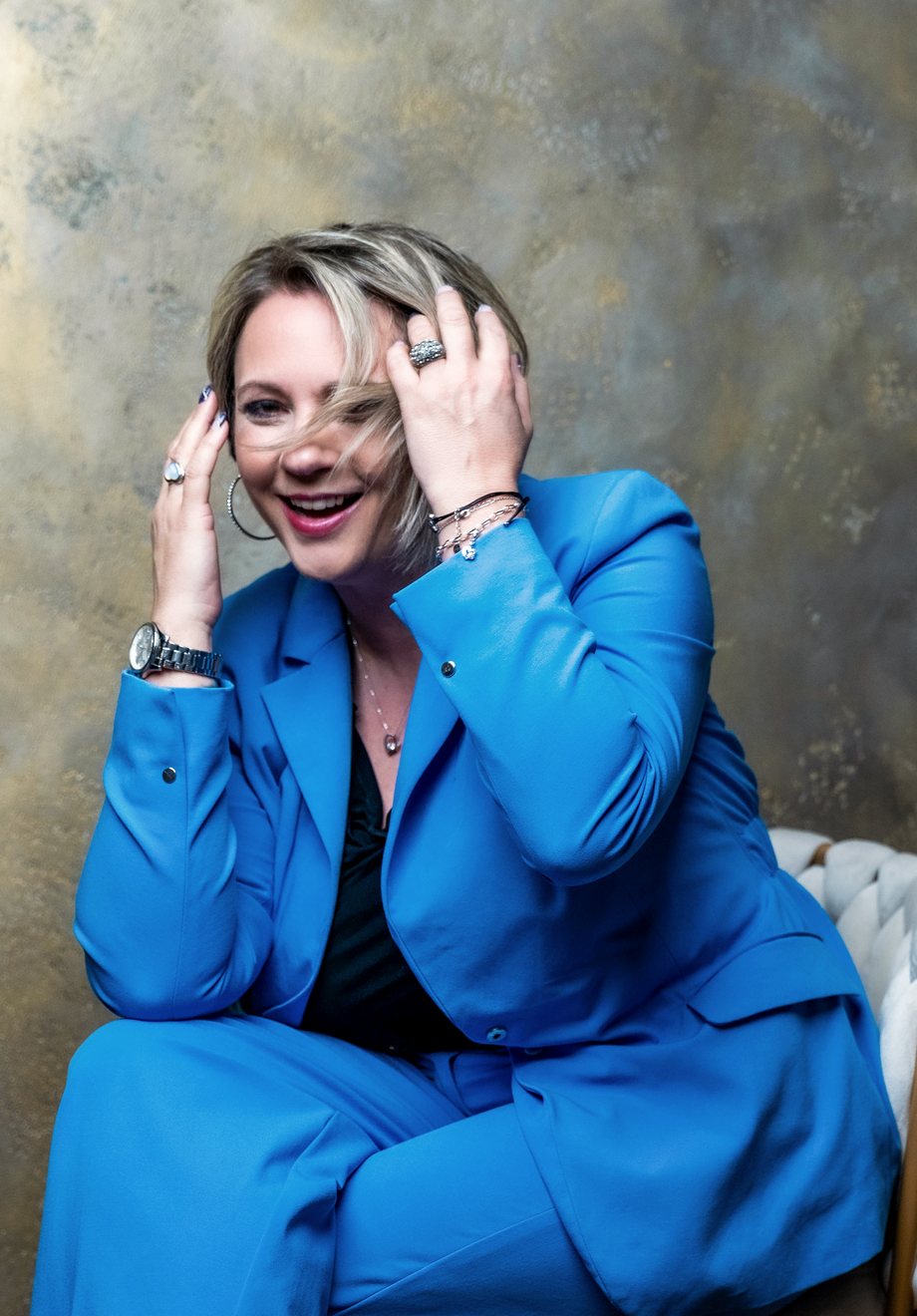 Angelika Stempfle im blauen Hosenanzug lacht, beide Hände im Haar